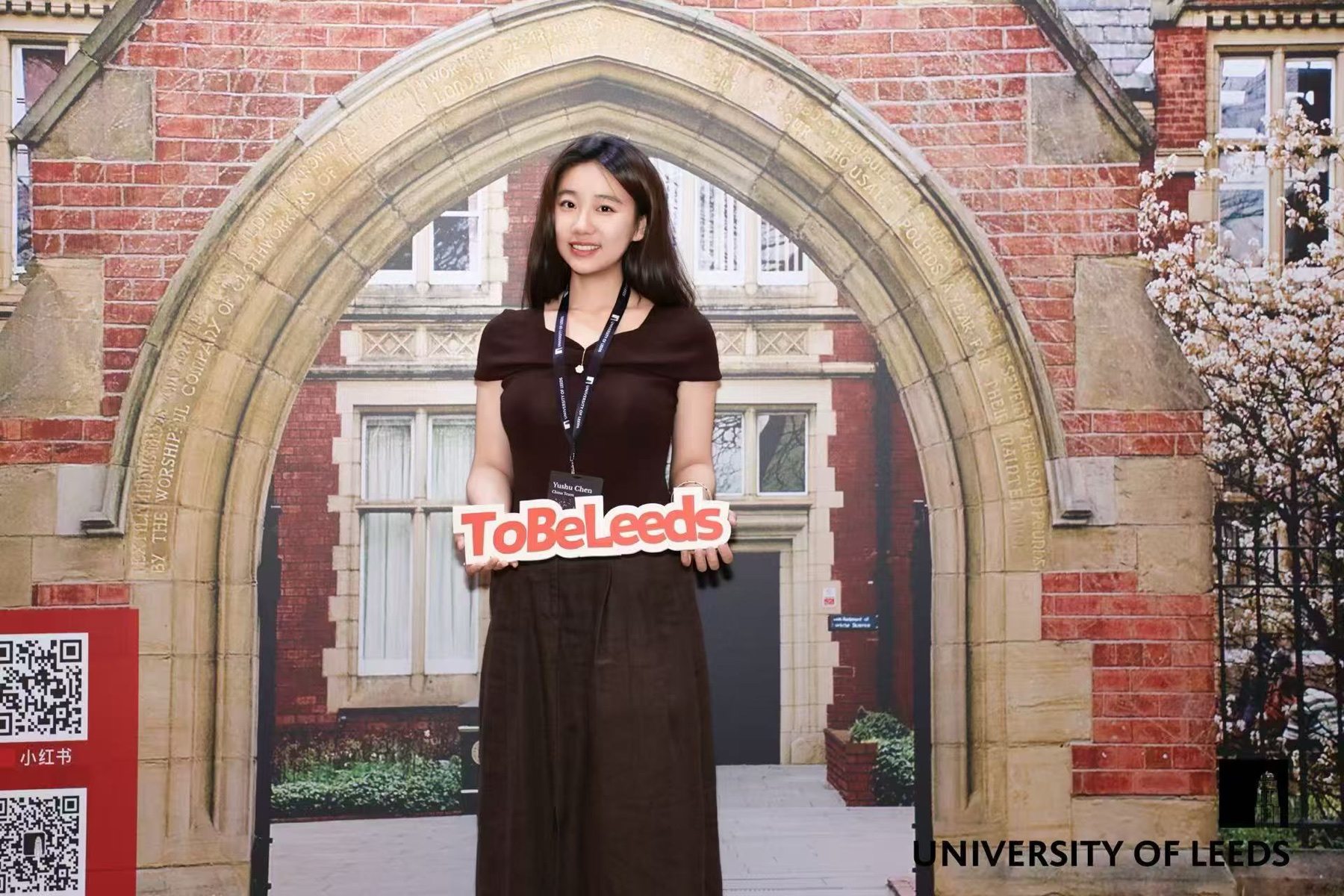Yushu Chen at a University of Leeds recruitment event in China, holding a ToBeLeeds sign in front of a printed campus backdrop, wearing a China Team name badge.